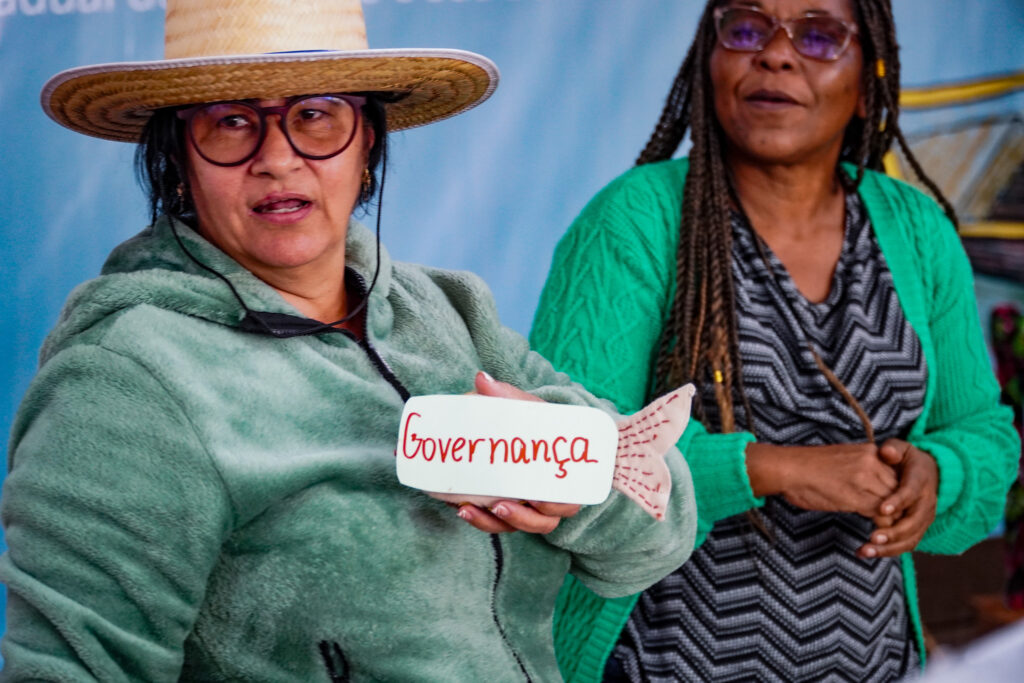 Pescadoras apresentam mística no início da reunião. Uma delas segura um peixe, com um papel colado com a palavra "Governança".