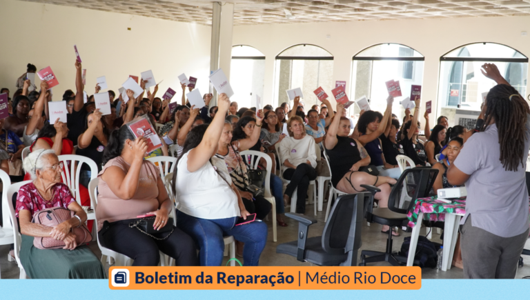 Um auditório de mulheres levantam um caderno de papel, que simbolizam o voto "sim" no Seminário Temático. Á frente, virada para elas, está uma mulher jovem, negra, empunhando um microfone e conferindo a quantidade de votos. Abaixo, os dizeres: Boletim da Reparação | Médio Rio Doce