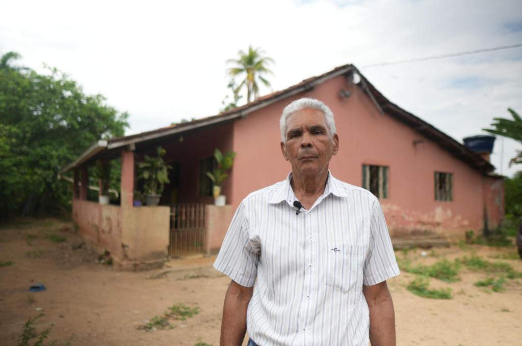 Geraldo Magela é um senhor grisalho, que olha para a câmera em frente a uma casa de cor salmão. Entre ele e a casa há um pátio descampado. O céu está nublado e acima da casa pode-se ver um palmeira bem alta.