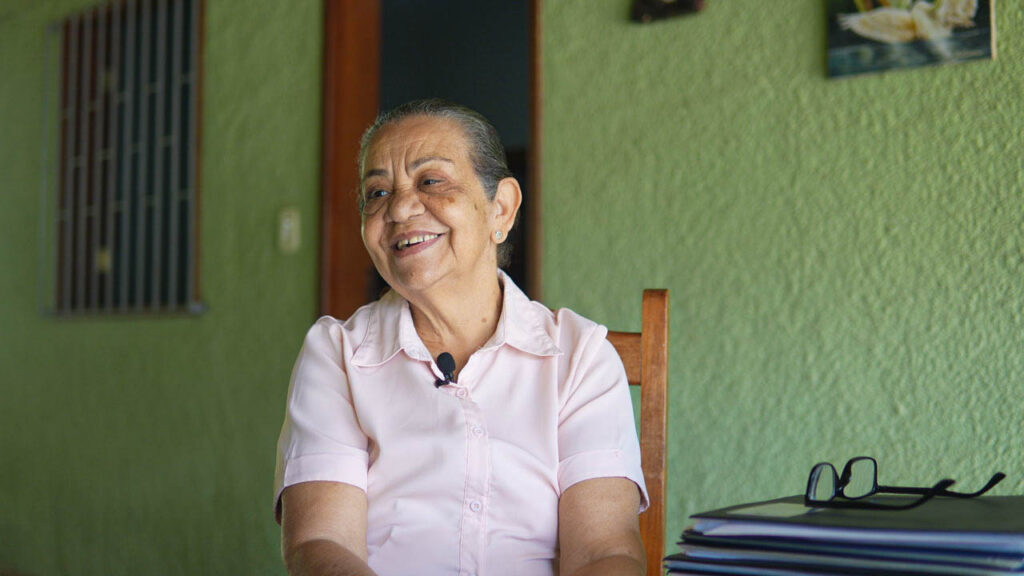 Drª. Graça está sentada na garagem de sua casa, com uma camisa rosa. Ela conversa com alguém fora do campo da imagem, e sorri. Tem cabelos levemente grisalhos e amarrados. Usa um brinco na orelha esquerda, e à sua frente há várias pastas com seus óculos repousando no topo. Ao fundo, uma janela e uma porta, com quadros aparecendo de relance na parede.